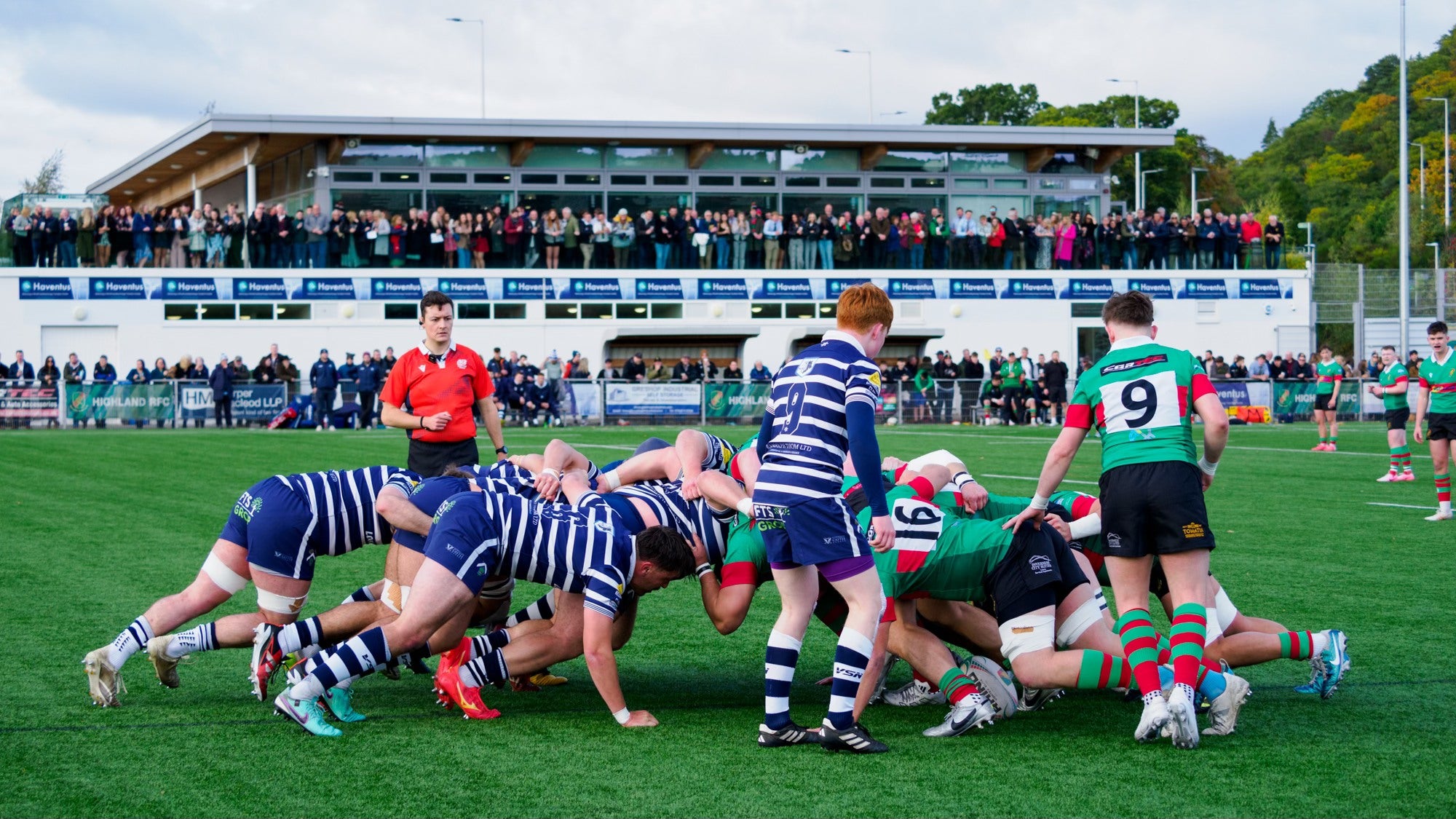 Rugby match in progress with players from two teams on a grass field, spectators in the stands.